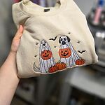 Sand ghost dogs with pumpkins sweatshirt held up by hand, stitched with custom coat colors: a cream dog and a black and white dog in ghost sheets