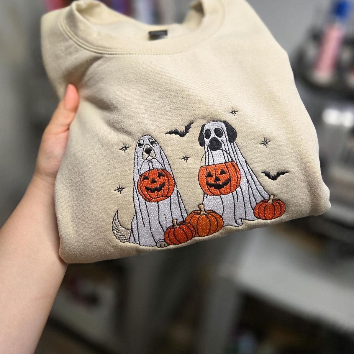 Ghost Dogs with Pumpkins Sweatshirt – Custom Coat Colors on Sand Sand ghost dogs with pumpkins sweatshirt held up by hand, stitched with custom coat colors: a cream dog and a black and white dog in ghost sheets
