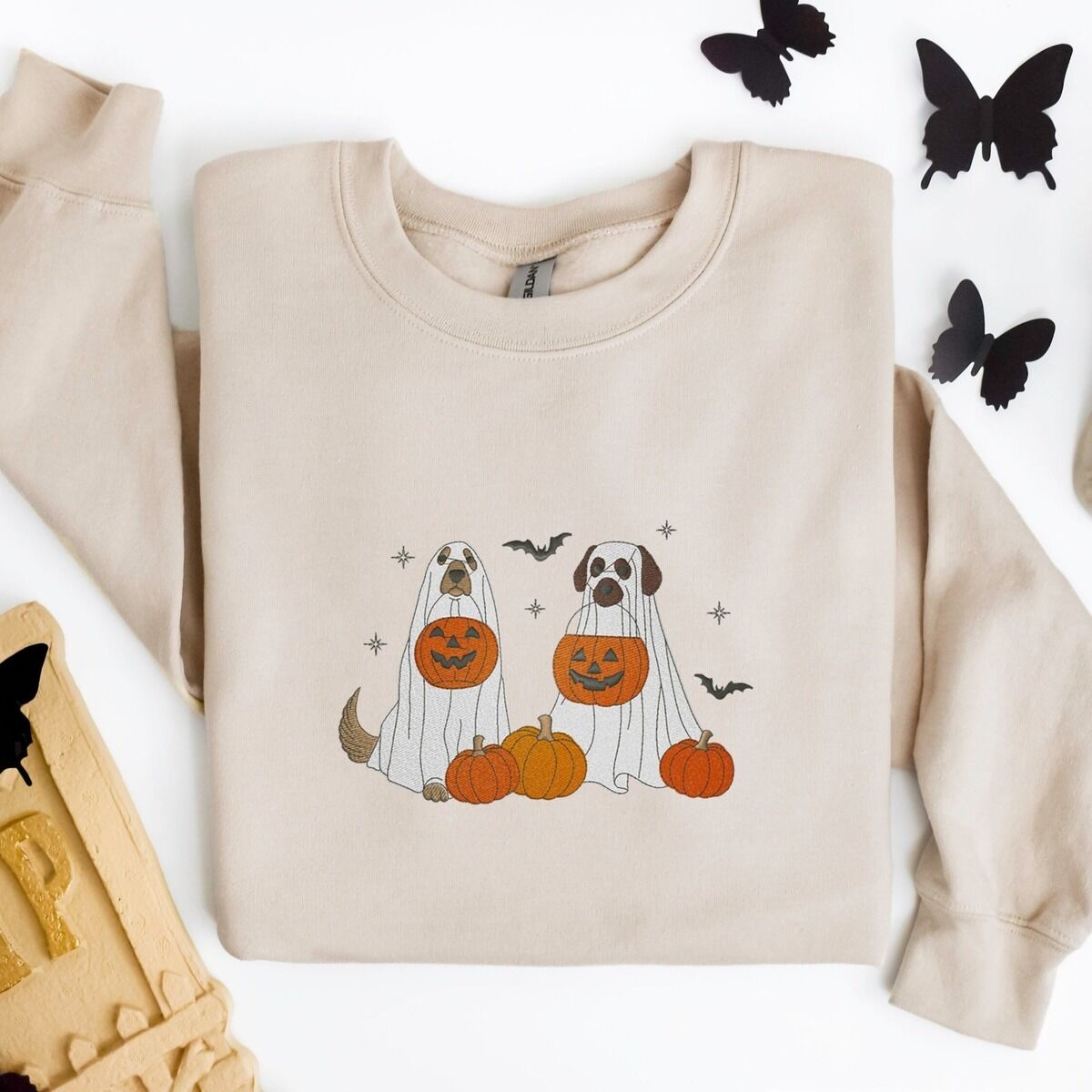 Ghost Dogs with Pumpkins Sweatshirt – Sand Sand crewneck sweatshirt laid flat with a skull, an RIP tombstone and black paper butterflies, embroidered with two dogs in white ghost sheets holding jack-o'-lantern buckets among pumpkins, with bats and stars