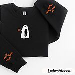 Black crewneck sweatshirt folded on a white background, embroidered with a white ghost hugging a black cat and holding a steaming jack-o'-lantern coffee mug, with small orange bats on the sleeves