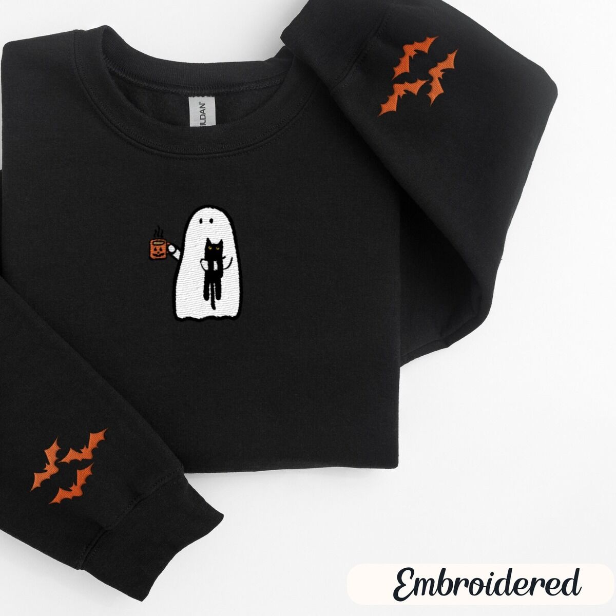 Ghost Drinking Coffee Sweatshirt – Black Black crewneck sweatshirt folded on a white background, embroidered with a white ghost hugging a black cat and holding a steaming jack-o'-lantern coffee mug, with small orange bats on the sleeves