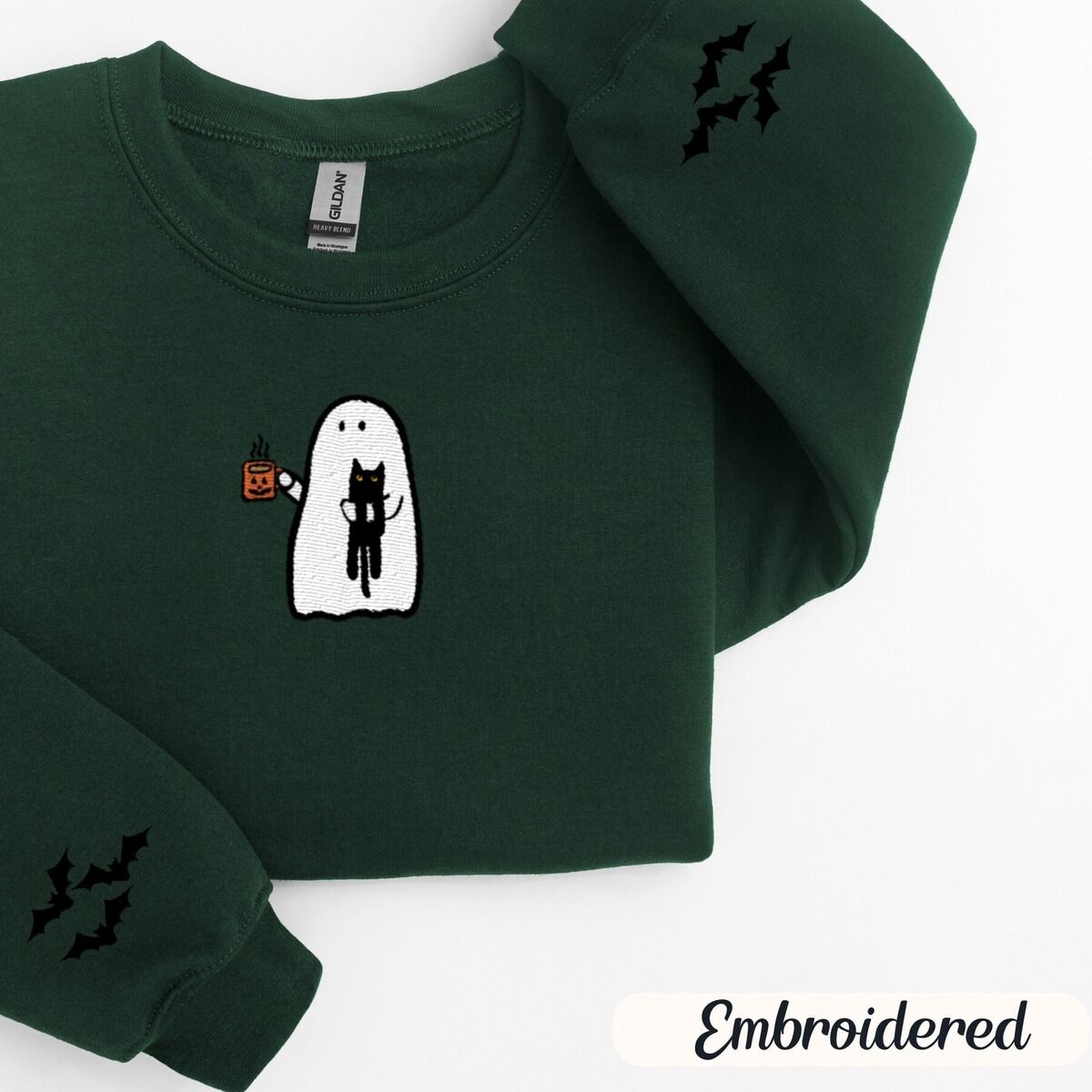 Ghost Drinking Coffee Sweatshirt – Forest Green Forest Green crewneck sweatshirt folded on a white background, embroidered with a white ghost hugging a black cat and holding a steaming jack-o'-lantern coffee mug, with small bats on the sleeves