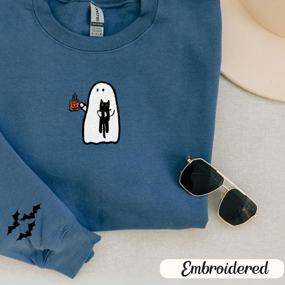 Ghost Drinking Coffee Sweatshirt – Indigo Blue Indigo Blue crewneck sweatshirt folded with a cream hat and sunglasses, embroidered with a white ghost hugging a black cat and holding a steaming jack-o'-lantern coffee mug, with small bats on the sleeves