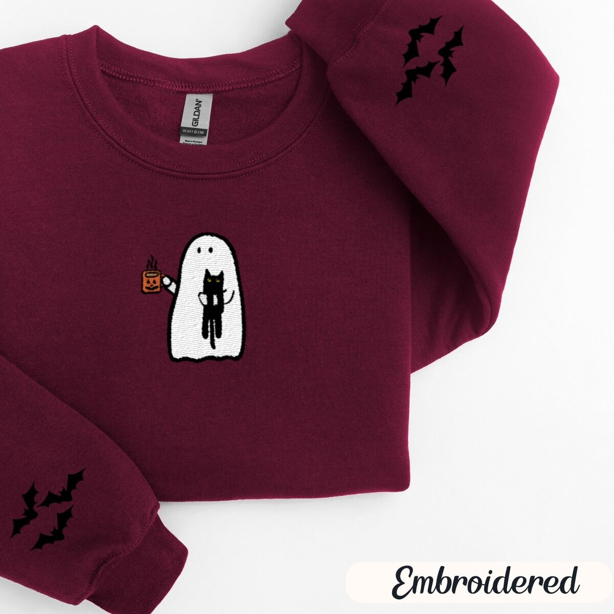 Ghost Drinking Coffee Sweatshirt – Maroon Maroon crewneck sweatshirt folded on a white background, embroidered with a white ghost hugging a black cat and holding a steaming jack-o'-lantern coffee mug, with small bats on the sleeves