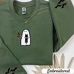 Military Green crewneck sweatshirt folded with a gold necklace and dried palm leaves, embroidered with a white ghost hugging a black cat and holding a steaming jack-o'-lantern coffee mug, with small bats on the sleeves