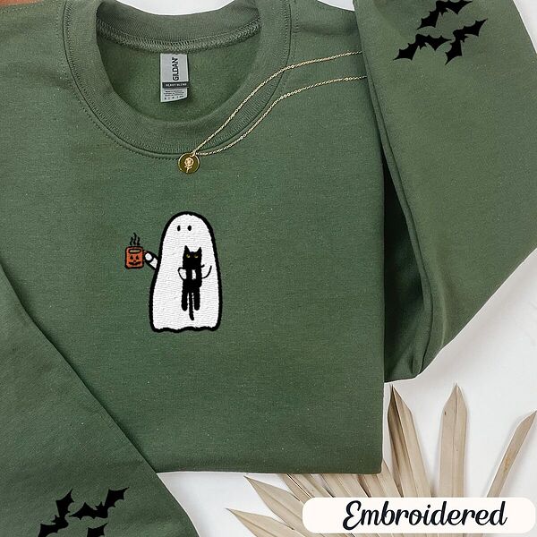 Military Green crewneck sweatshirt folded with a gold necklace and dried palm leaves, embroidered with a white ghost hugging a black cat and holding a steaming jack-o'-lantern coffee mug, with small bats on the sleeves