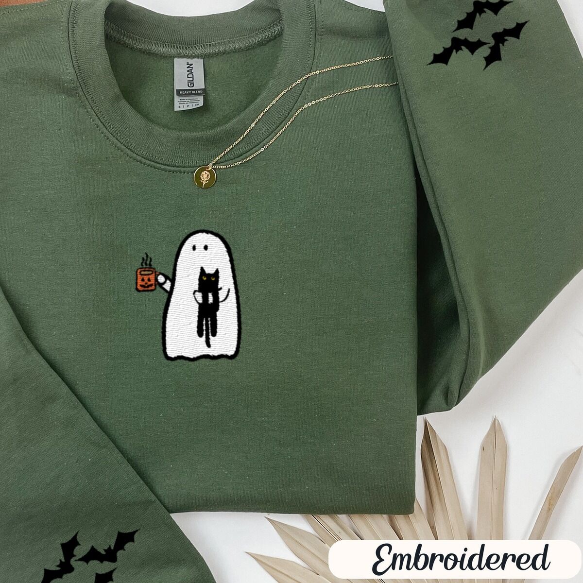 Ghost Drinking Coffee Sweatshirt – Military Green Military Green crewneck sweatshirt folded with a gold necklace and dried palm leaves, embroidered with a white ghost hugging a black cat and holding a steaming jack-o'-lantern coffee mug, with small bats on the sleeves