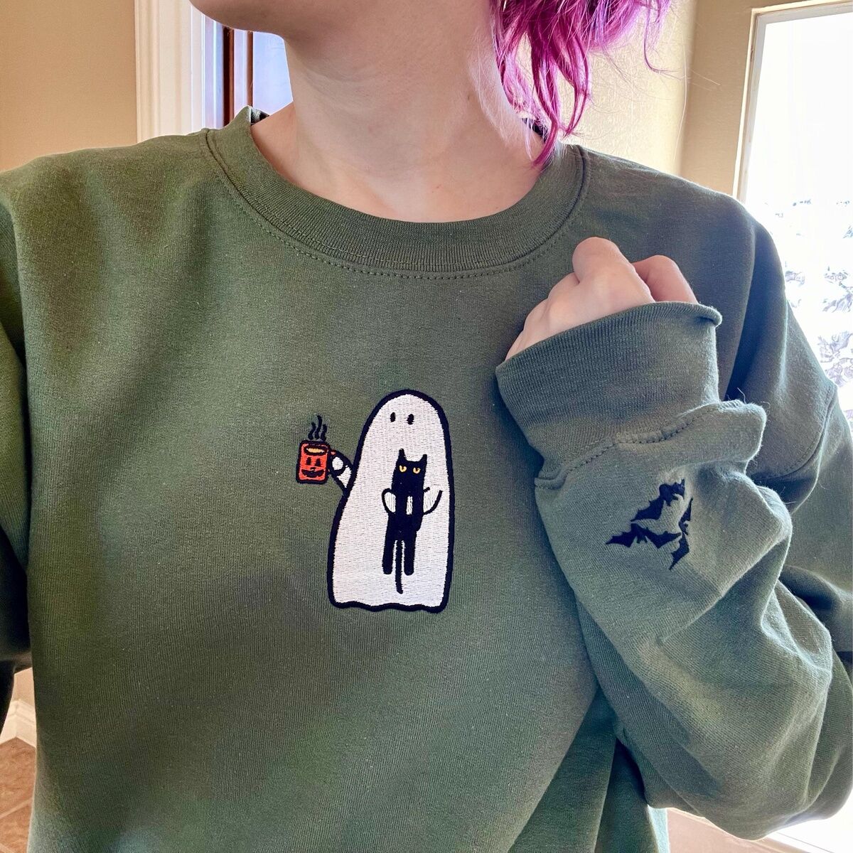 Ghost Drinking Coffee Sweatshirt – Customer Photo 1 Customer photo: close-up of a Military Green ghost drinking coffee sweatshirt being worn, with the embroidered ghost holding a black cat and a jack-o'-lantern mug, and bats on the sleeve