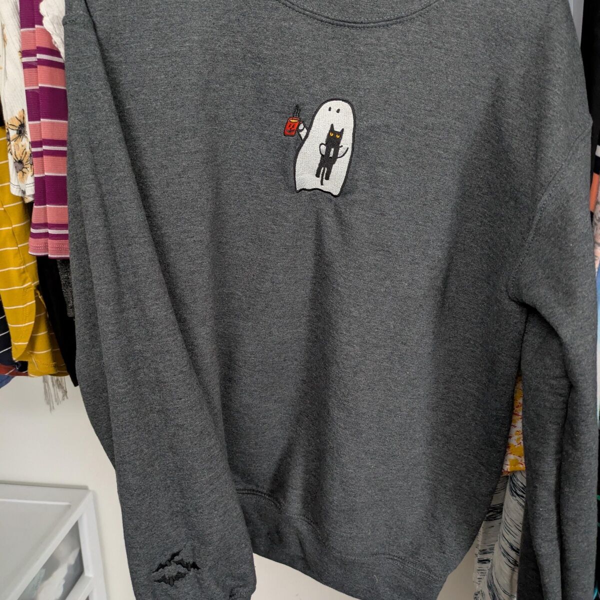 Ghost Drinking Coffee Sweatshirt – Customer Photo 3 Customer photo: dark gray ghost drinking coffee sweatshirt on a hanger, with the embroidered ghost and black cat on the chest and bats near the cuff