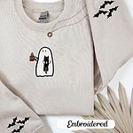 Sand crewneck sweatshirt folded with a gold necklace and dried palm leaves, embroidered with a white ghost hugging a black cat and holding a steaming jack-o'-lantern coffee mug, with small bats on the sleeves