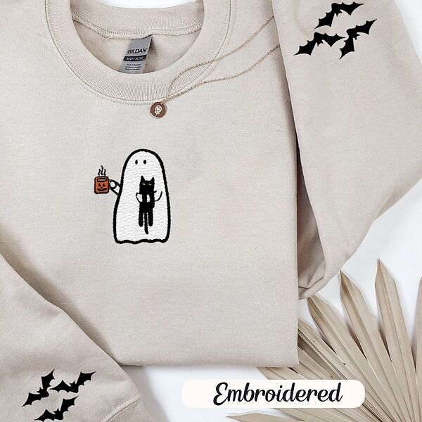 Sand crewneck sweatshirt folded with a gold necklace and dried palm leaves, embroidered with a white ghost hugging a black cat and holding a steaming jack-o'-lantern coffee mug, with small bats on the sleeves