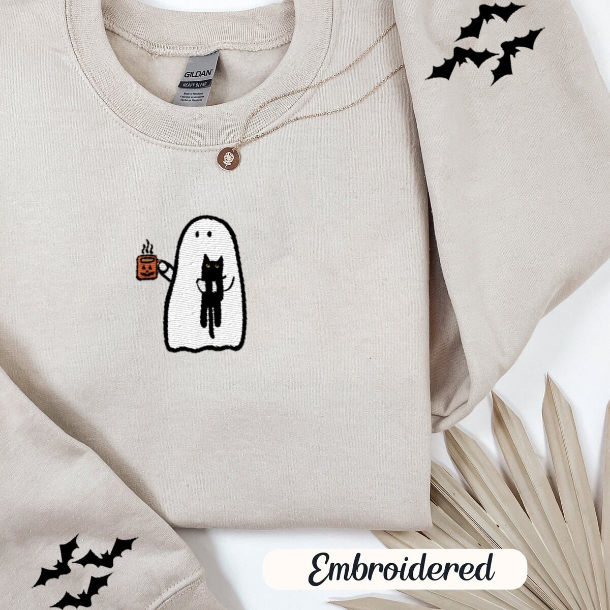 Ghost Drinking Coffee Sweatshirt – Sand Sand crewneck sweatshirt folded with a gold necklace and dried palm leaves, embroidered with a white ghost hugging a black cat and holding a steaming jack-o'-lantern coffee mug, with small bats on the sleeves