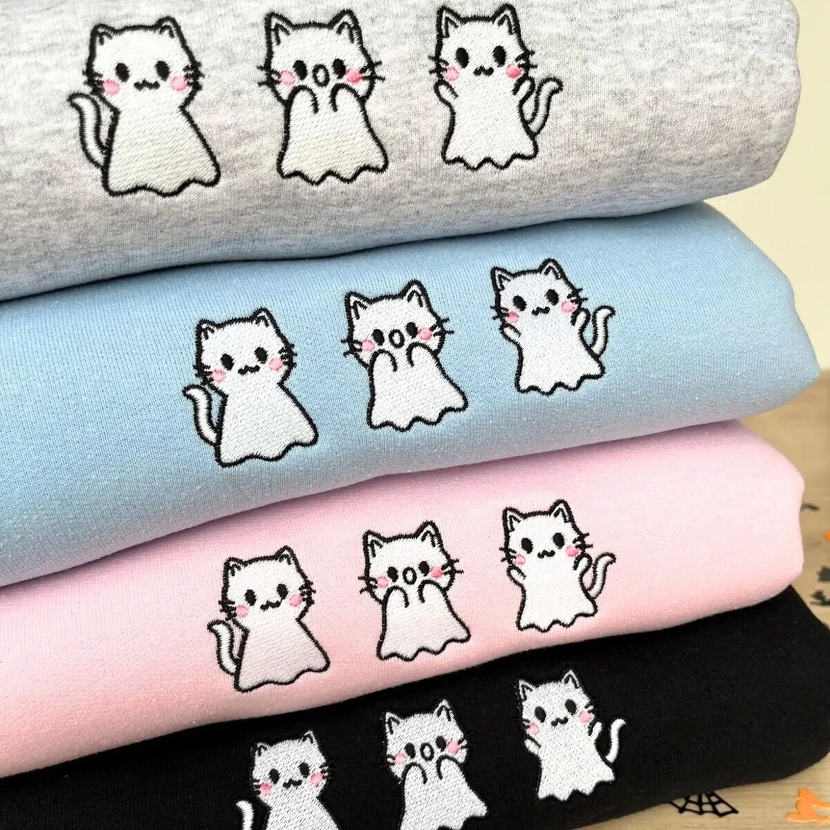 Ghost Kitten Embroidery – Real Stitching Real embroidery of three white ghost kittens with pink cheeks, stitched on four stacked crewneck sweatshirts in heather grey, light blue, light pink and black
