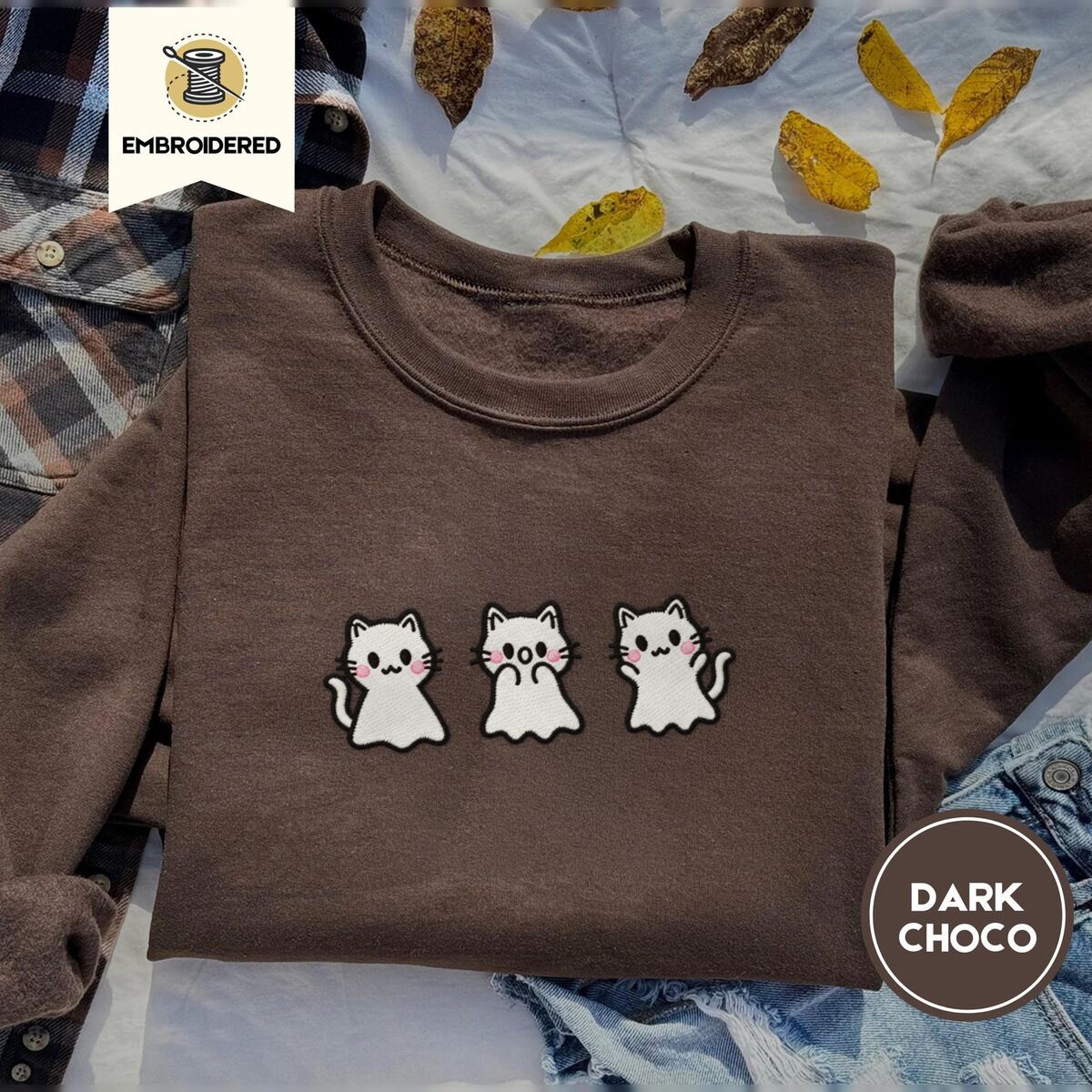 Ghost Kitten Sweatshirt – Dark Chocolate Dark Chocolate crewneck sweatshirt on a plaid flannel shirt with yellow autumn leaves and denim shorts, embroidered with three white ghost kittens with pink cheeks and black outlines