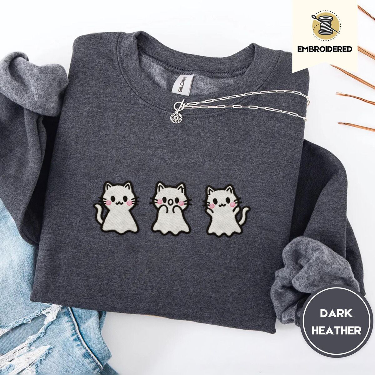 Ghost Kitten Sweatshirt – Dark Heather Dark Heather crewneck sweatshirt folded with ripped jeans and a silver chain necklace, embroidered with three white ghost kittens with pink cheeks and black outlines
