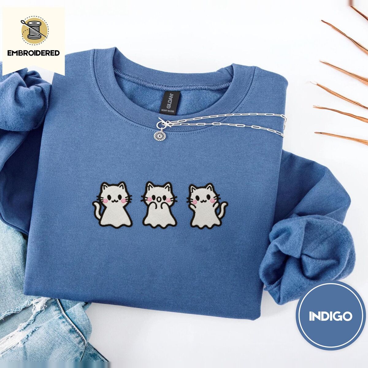 Ghost Kitten Sweatshirt – Indigo Blue Indigo Blue crewneck sweatshirt folded with light jeans and dried twigs, embroidered with three white ghost kittens with pink cheeks and black outlines