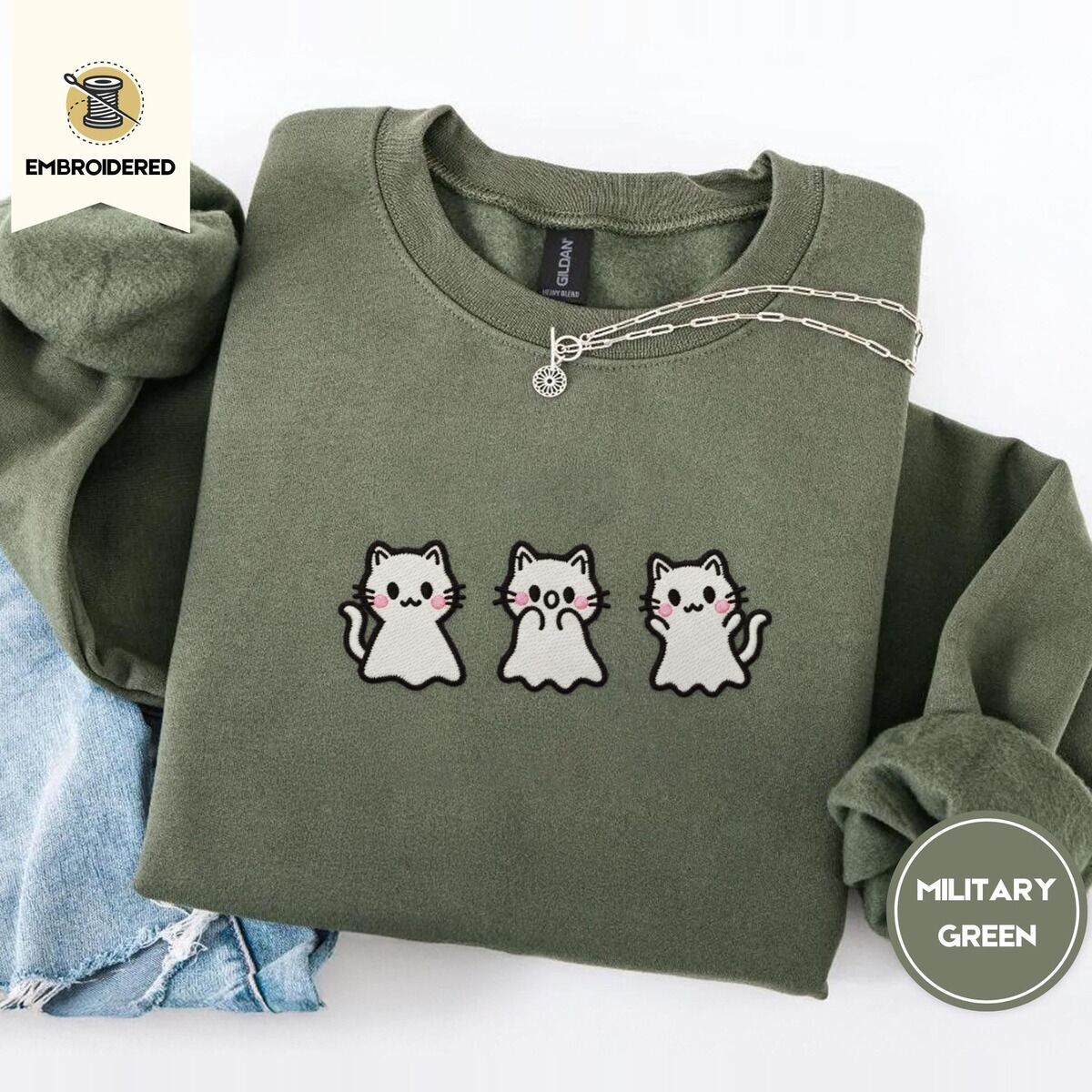 Ghost Kitten Sweatshirt – Military Green Military Green crewneck sweatshirt folded with light jeans and a silver chain necklace, embroidered with three white ghost kittens with pink cheeks and black outlines