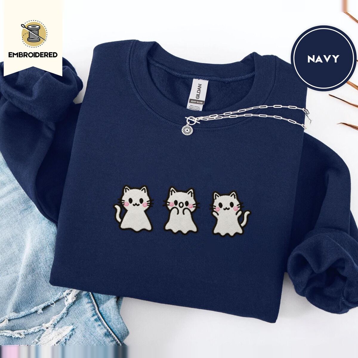 Ghost Kitten Sweatshirt – Navy Navy crewneck sweatshirt folded with ripped jeans and a silver chain necklace, embroidered with three white ghost kittens with pink cheeks and black outlines