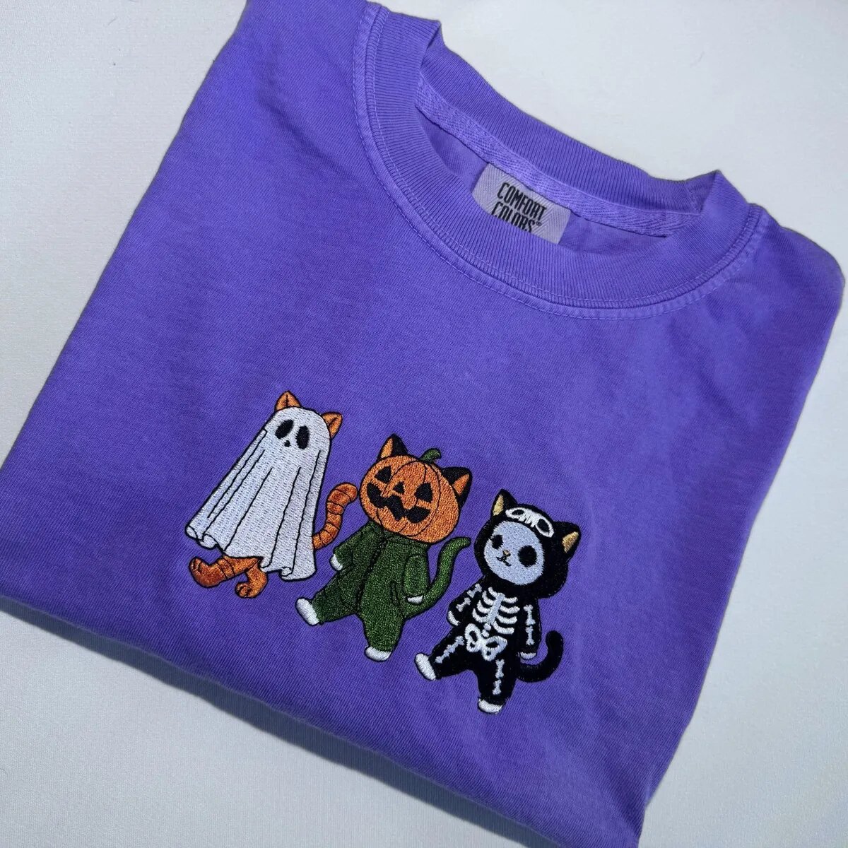 Ghost, Pumpkin and Skeleton Cats Embroidered Tee Angled view of the ghost cat, pumpkin cat and skeleton cat embroidery on a violet T-shirt