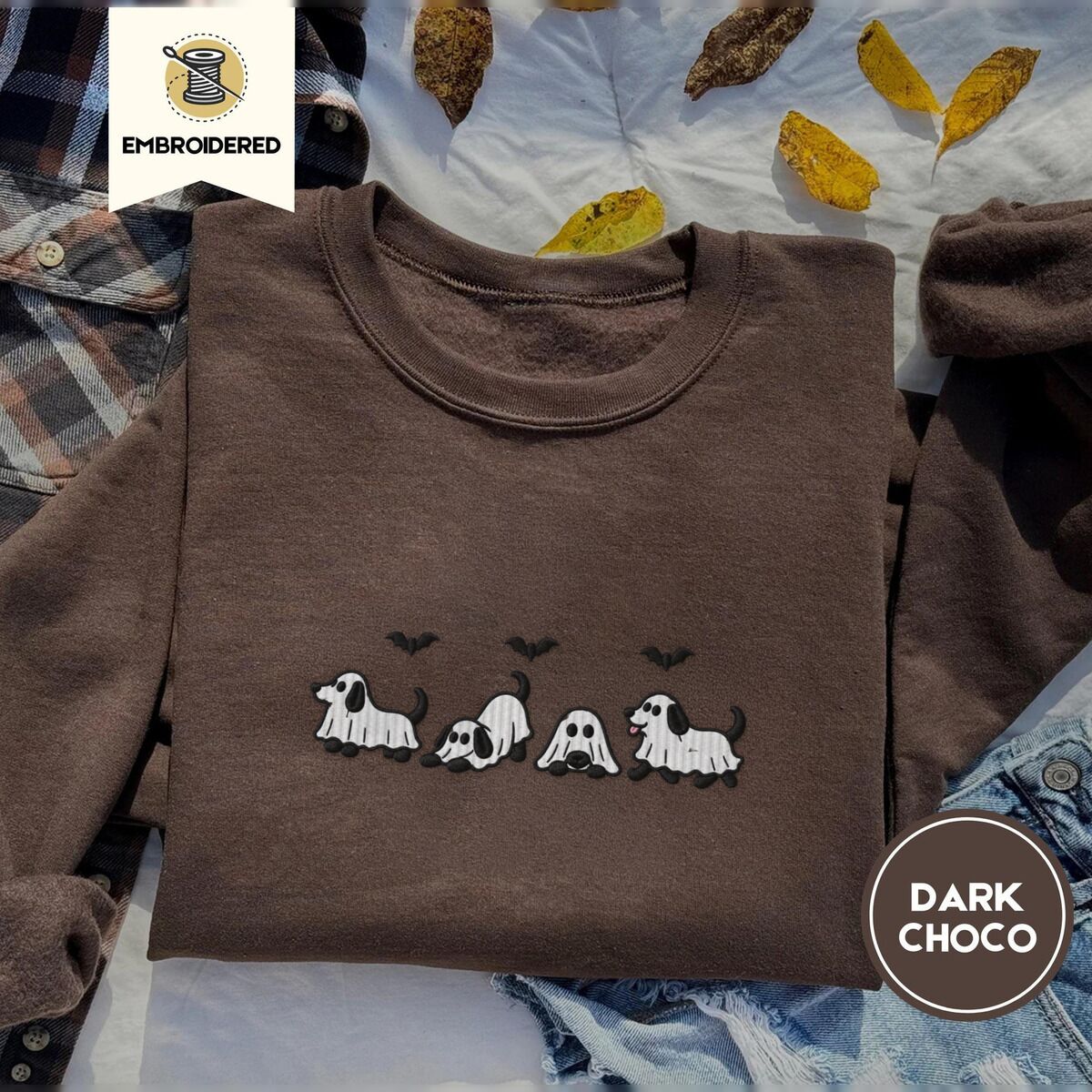 Ghost Puppy Sweatshirt – Dark Chocolate Dark Chocolate crewneck sweatshirt on a plaid flannel shirt with yellow autumn leaves and denim shorts, embroidered with four puppies in white ghost sheets with black floppy ears and tails under three flying black bats