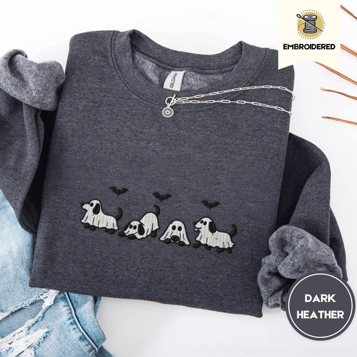Ghost Puppy Sweatshirt – Dark Heather Dark Heather crewneck sweatshirt folded with ripped jeans and a silver chain necklace, embroidered with four puppies in white ghost sheets with black floppy ears and tails under three flying black bats