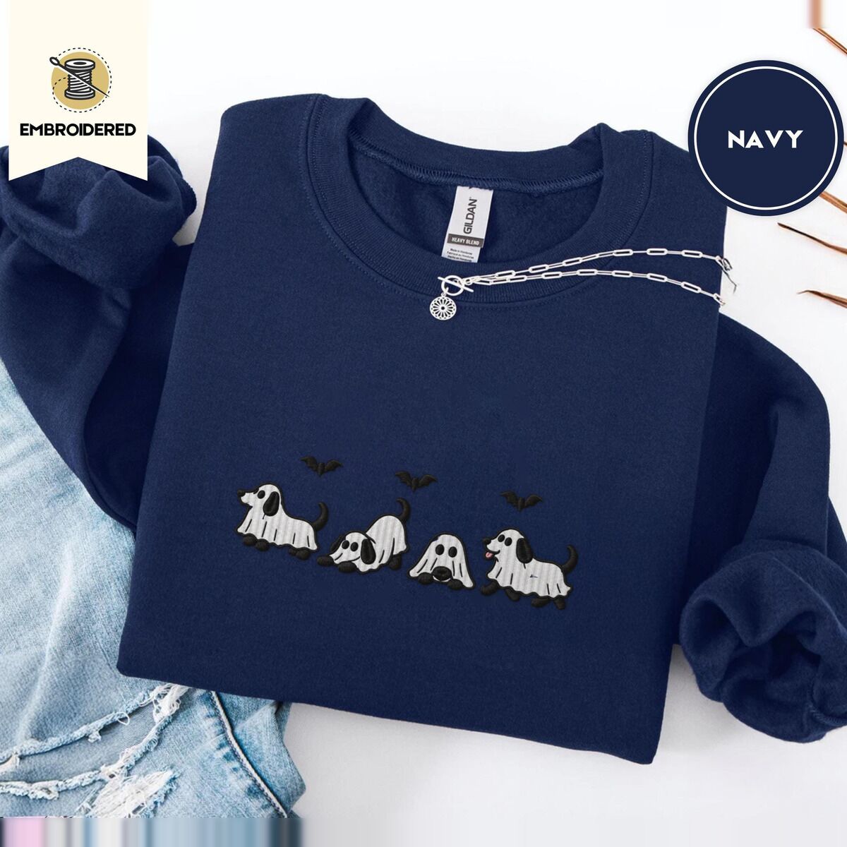Ghost Puppy Sweatshirt – Navy Navy crewneck sweatshirt folded with ripped jeans and a silver chain necklace, embroidered with four puppies in white ghost sheets with black floppy ears and tails under three flying black bats