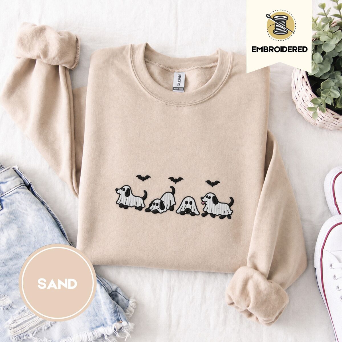 Ghost Puppy Sweatshirt – Sand Sand crewneck sweatshirt folded with denim shorts, white sneakers and a potted plant, embroidered with four puppies in white ghost sheets with black floppy ears and tails under three flying black bats