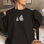 Woman in a Black crewneck sweatshirt embroidered with a ghost sipping iced coffee and carrying a jack-o'-lantern pail while walking a little yellow lab in a ghost sheet on a leash