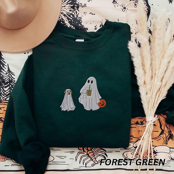 Forest Green crewneck sweatshirt laid flat with a brown hat and pampas grass, embroidered with a ghost sipping iced coffee and carrying a jack-o'-lantern pail while walking a little yellow lab in a ghost sheet on a leash