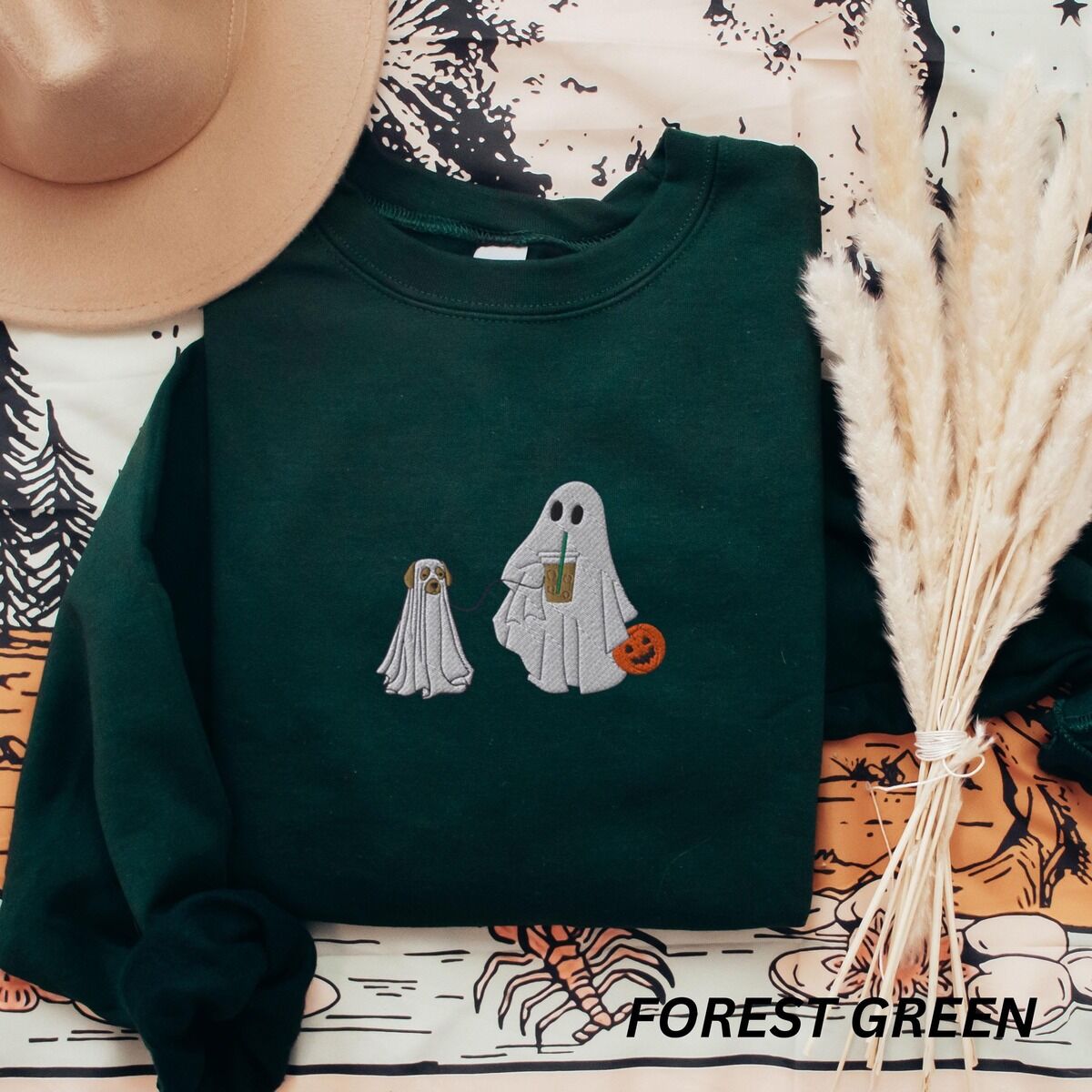 Ghost Walking Dog Sweatshirt – Forest Green Forest Green crewneck sweatshirt laid flat with a brown hat and pampas grass, embroidered with a ghost sipping iced coffee and carrying a jack-o'-lantern pail while walking a little yellow lab in a ghost sheet on a leash
