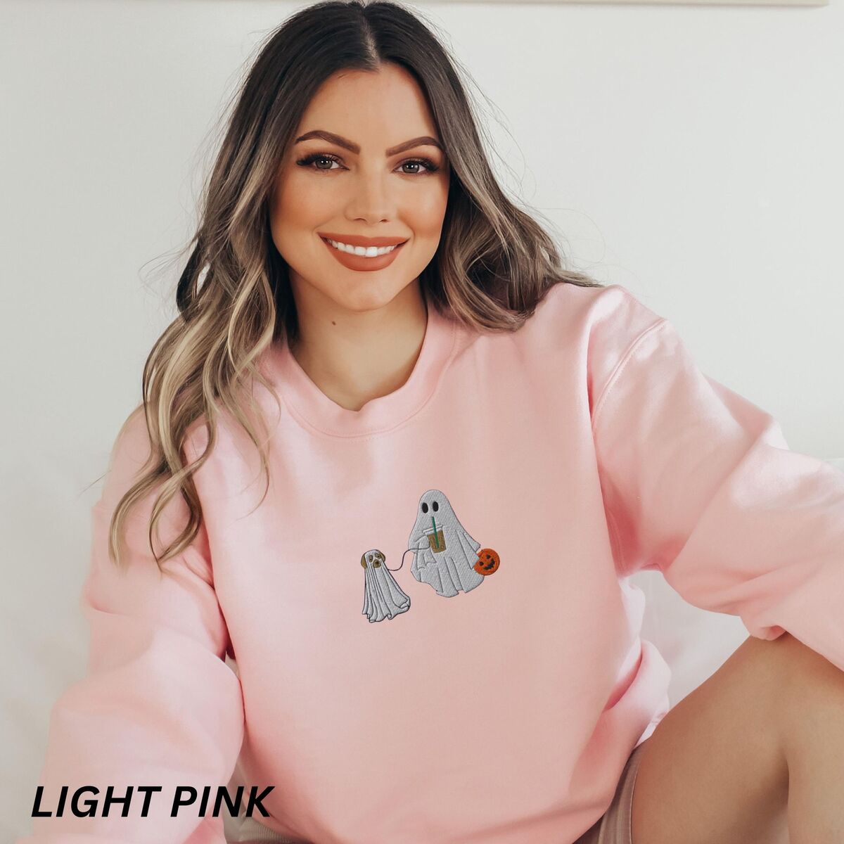 Ghost Walking Dog Sweatshirt - Light Pink Smiling woman sitting in a Light Pink crewneck sweatshirt embroidered with a ghost sipping iced coffee and carrying a jack-o'-lantern pail while walking a little yellow lab in a ghost sheet on a leash