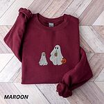 Maroon crewneck sweatshirt laid flat on a wood floor, embroidered with a ghost sipping iced coffee and carrying a jack-o'-lantern pail while walking a little yellow lab in a ghost sheet on a leash