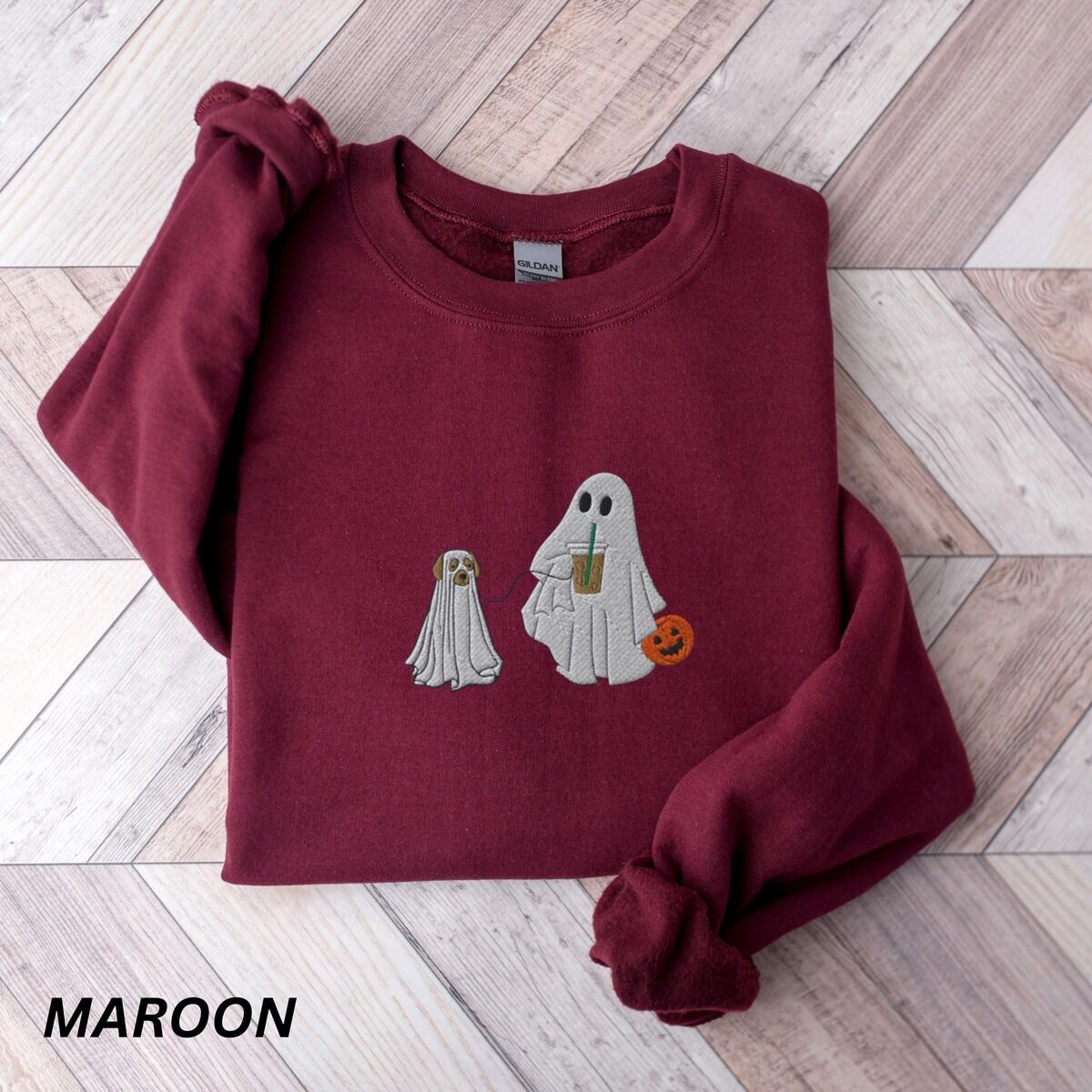 Ghost Walking Dog Sweatshirt – Maroon Maroon crewneck sweatshirt laid flat on a wood floor, embroidered with a ghost sipping iced coffee and carrying a jack-o'-lantern pail while walking a little yellow lab in a ghost sheet on a leash