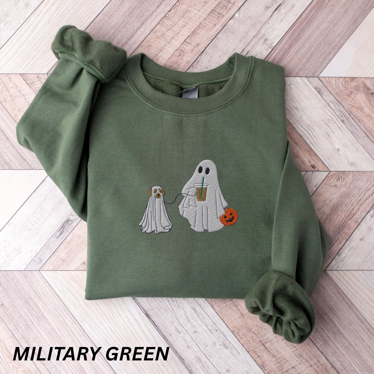 Ghost Walking Dog Sweatshirt – Military Green Military Green crewneck sweatshirt laid flat on a wood floor, embroidered with a ghost sipping iced coffee and carrying a jack-o'-lantern pail while walking a little yellow lab in a ghost sheet on a leash