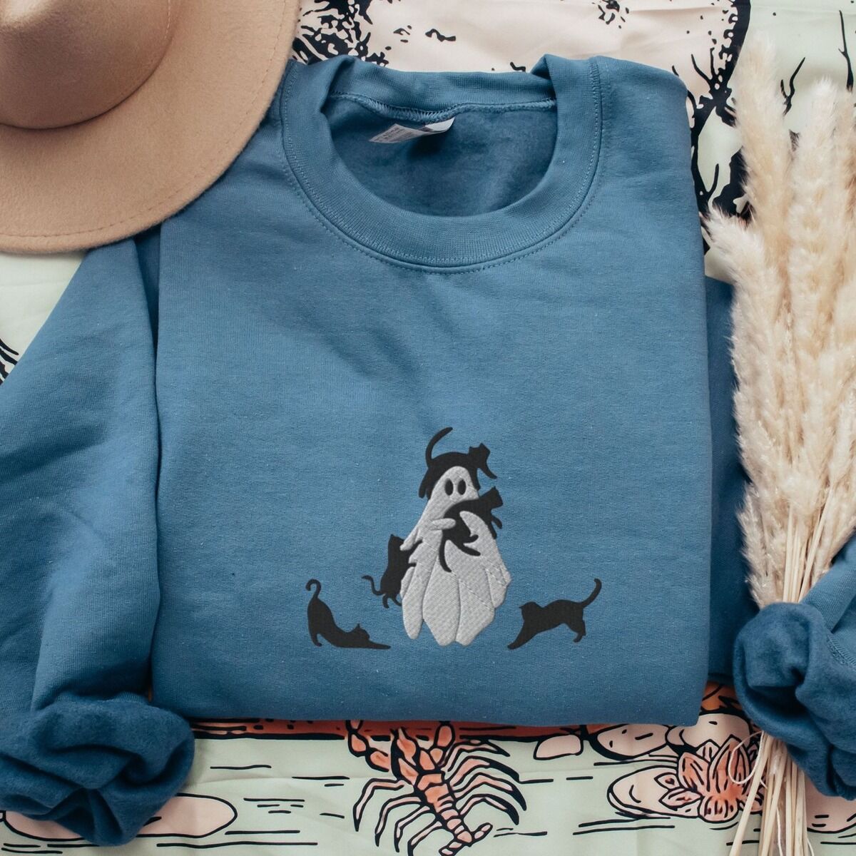 Ghost with Black Cats Sweatshirt – Indigo Blue Indigo Blue crewneck sweatshirt folded with a tan felt hat and pampas grass, embroidered with a ghost covered in black cats, one draped over its head, one in its arms, one climbing its side and two stretching at its feet