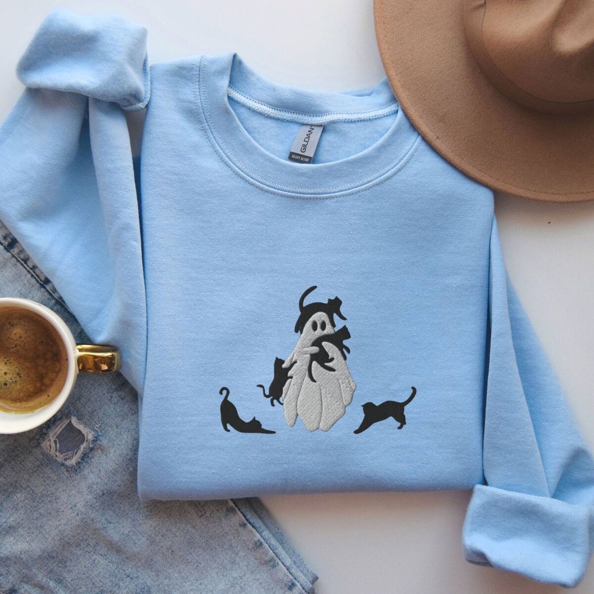 Ghost with Black Cats Sweatshirt – Light Blue Light Blue crewneck sweatshirt folded with jeans, a tan felt hat and a cup of coffee, embroidered with a ghost covered in black cats, one draped over its head, one in its arms, one climbing its side and two stretching at its feet