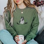 Woman holding a mug in a Military Green crewneck sweatshirt embroidered with a ghost covered in black cats, one draped over its head, one in its arms, one climbing its side and two stretching at its feet