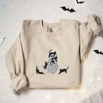 Sand crewneck sweatshirt laid flat with paper bats and fairy lights, embroidered with a ghost covered in black cats, one draped over its head, one in its arms, one climbing its side and two stretching at its feet