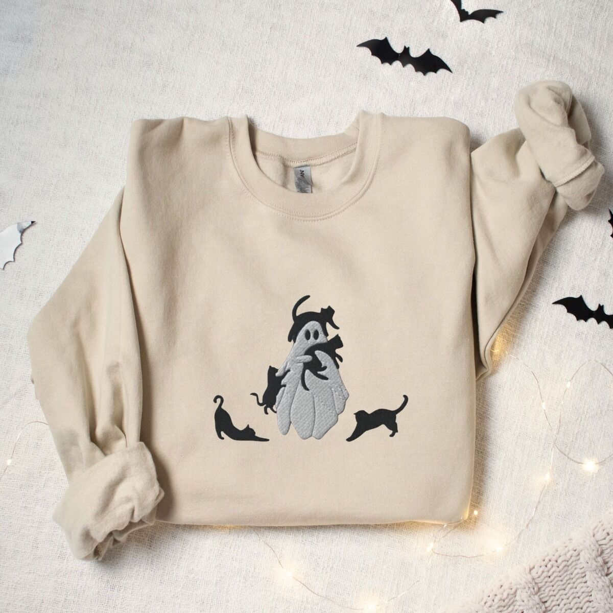 Ghost with Black Cats Sweatshirt – Sand Sand crewneck sweatshirt laid flat with paper bats and fairy lights, embroidered with a ghost covered in black cats, one draped over its head, one in its arms, one climbing its side and two stretching at its feet