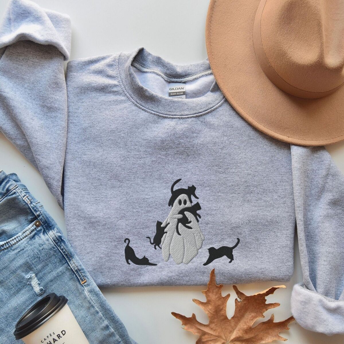 Ghost with Black Cats Sweatshirt – Sport Grey Sport Grey crewneck sweatshirt folded with jeans, a tan felt hat, an autumn leaf and a coffee cup, embroidered with a ghost covered in black cats, one draped over its head, one in its arms, one climbing its side and two stretching at its feet