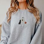 Woman with long hair in a grey crewneck sweatshirt embroidered with a ghost lab mom sipping iced coffee and carrying a jack-o'-lantern pail, with a black lab sitting against her