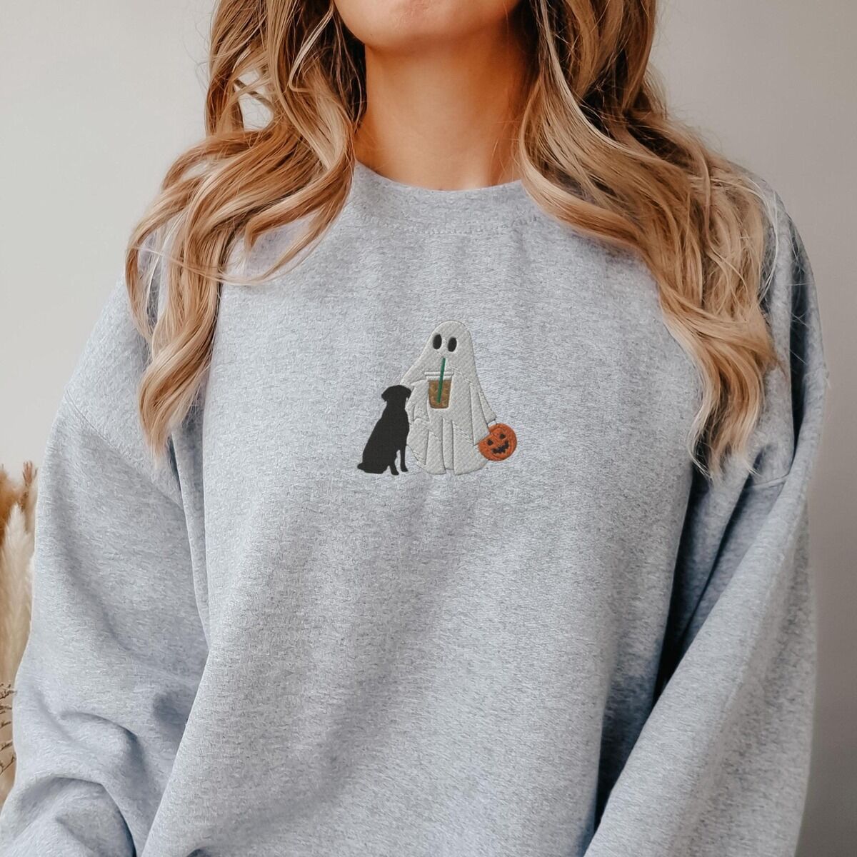 Ghost with Black Lab Sweatshirt - worn in grey Woman with long hair in a grey crewneck sweatshirt embroidered with a ghost lab mom sipping iced coffee and carrying a jack-o'-lantern pail, with a black lab sitting against her