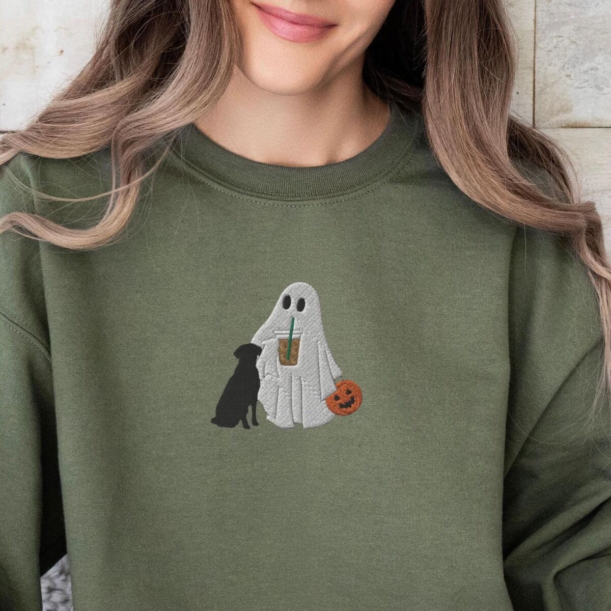 Ghost with Black Lab Sweatshirt - worn Close-up of a woman in a green crewneck sweatshirt embroidered with a ghost lab mom sipping iced coffee and carrying a jack-o'-lantern pail, with a black lab sitting against her