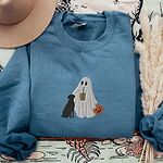 Indigo Blue crewneck sweatshirt laid flat with a brown hat and pampas grass, embroidered with a ghost lab mom sipping iced coffee and carrying a jack-o'-lantern pail, with a black lab sitting against her