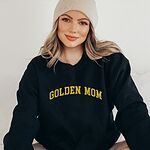 Woman in a beanie and a Black crewneck sweatshirt embroidered with GOLDEN MOM in arched mustard gold varsity letters