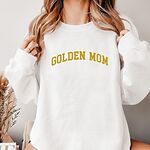 Woman in a White crewneck sweatshirt embroidered with GOLDEN MOM in arched mustard gold varsity letters