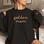 Woman in a Black crewneck sweatshirt embroidered with golden mom in gold script and a golden retriever silhouette on the cuff