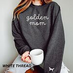 Woman holding a mug in a Dark Heather crewneck sweatshirt embroidered with golden mom in white script and a white golden retriever silhouette on the cuff