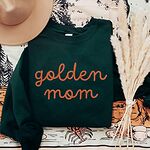 Forest Green crewneck sweatshirt laid flat with a hat and dried pampas, embroidered with golden mom in rust script across the chest