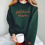 Woman holding a mug in a Forest Green crewneck sweatshirt embroidered with golden mom in rust script and a rust golden retriever silhouette on the cuff